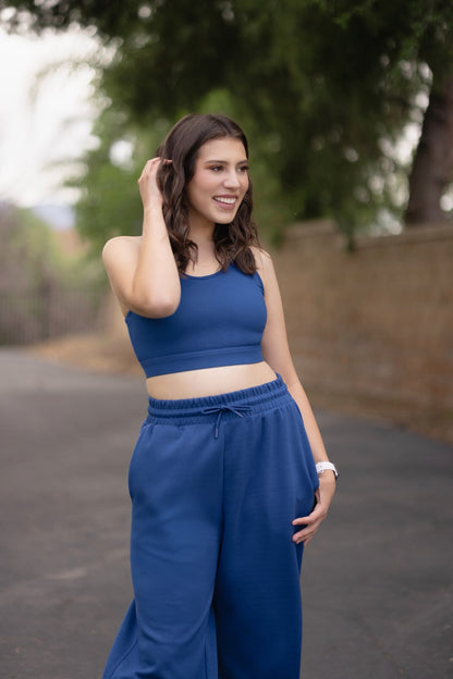 Woman wearing a solid peony blue LuLaRoe Rise Invincible sports bra with racerback silhouette and matching blue athleisure pants, styled outdoors with a clean, modern activewear look with trees in the background