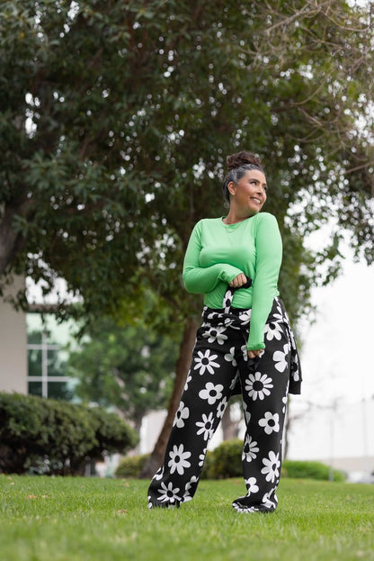 Model wearing LuLaRoe RISE Recharge Pull-On Sweatpant in Midnight Mod Daisy black fleece with oversized white retro daisy print styled outdoors on grass with relaxed wide-leg fit standing near trees and a building in the background.
