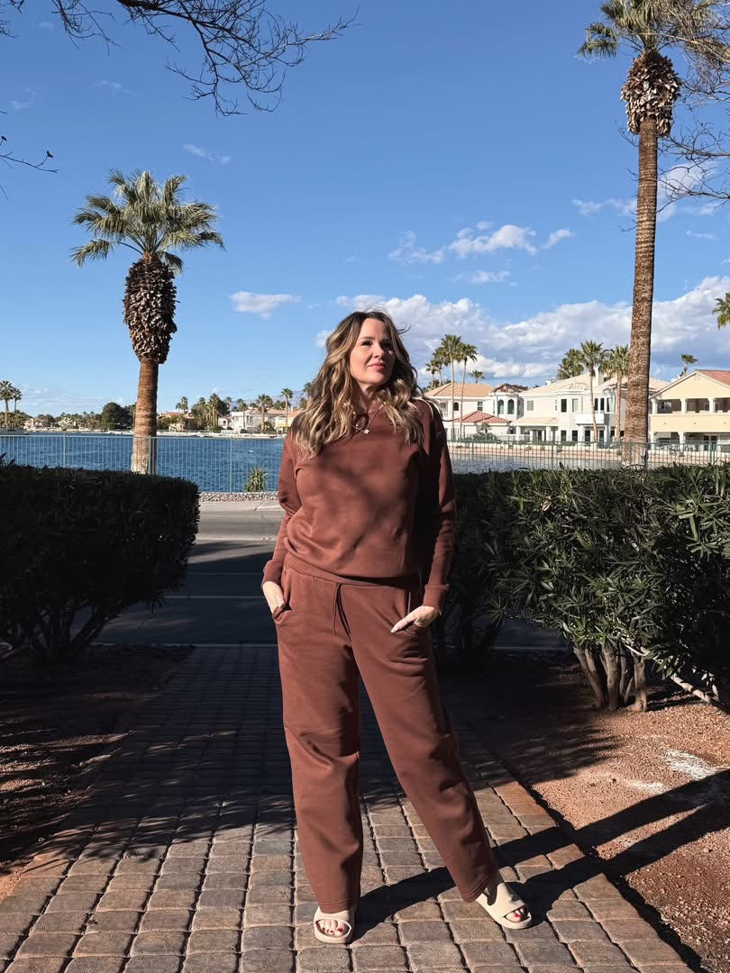 Model wearing LuLaRoe RISE Recharge Pull-On Sweatpant in Solid Cocoa Ember warm brown fleece with relaxed wide-leg silhouette styled outdoors near palm trees and lake with slide sandals