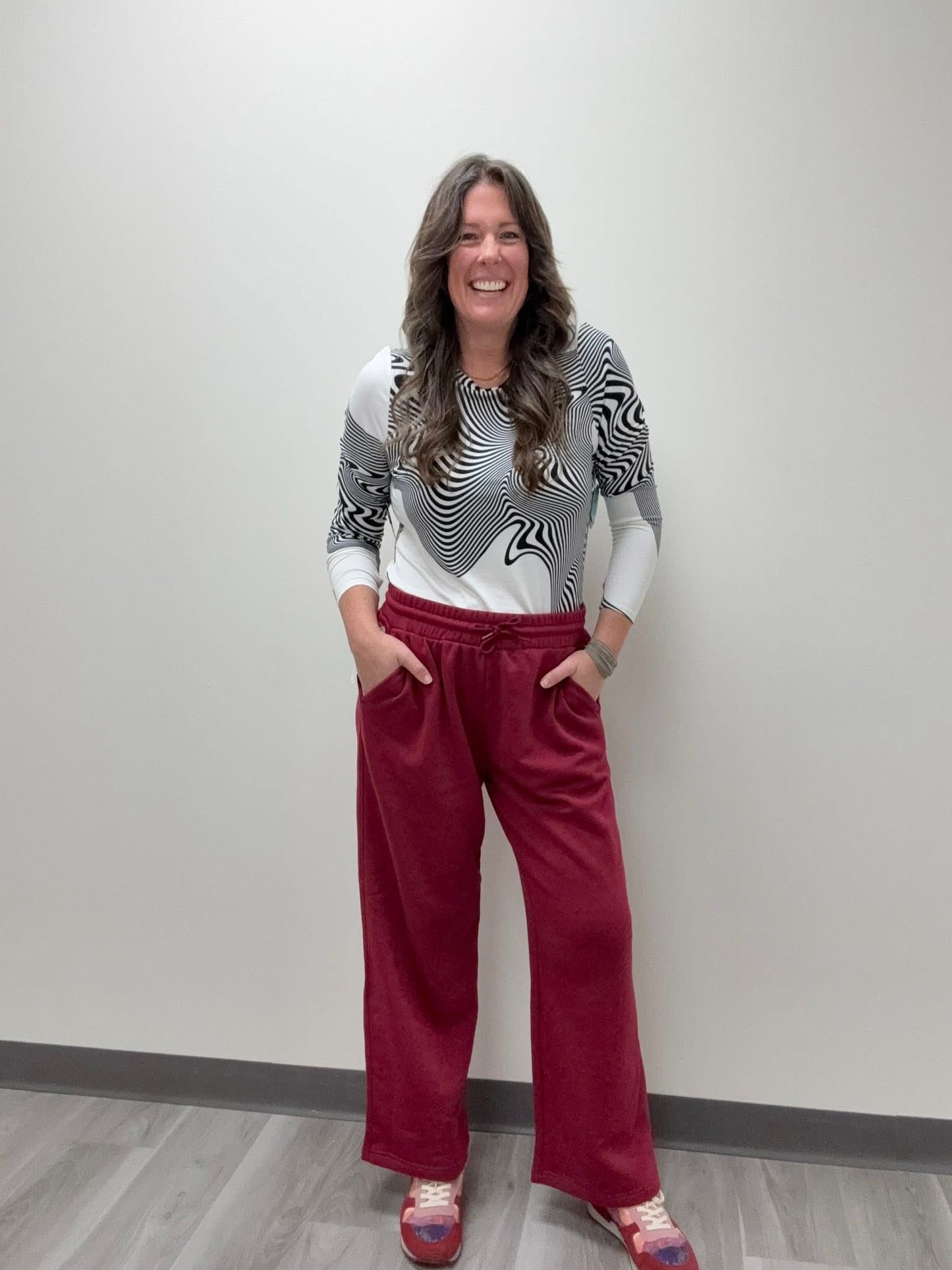LuLaRoe RISE Recharge Pull-On Sweatpant in Solid Crimson Reserve deep red fleece with relaxed wide-leg fit and elastic drawstring waist, styled with black and white abstract top against a plain wall.