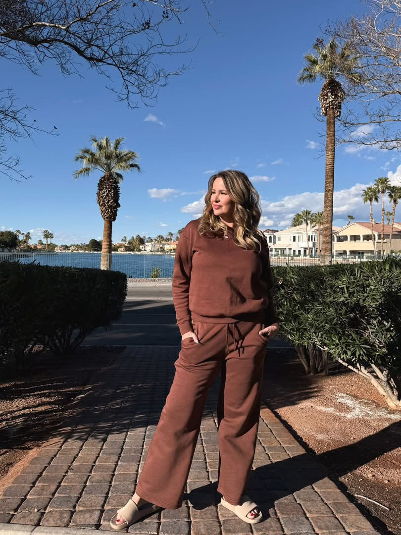 Woman wearing LuLaRoe RISE Reset fleece crew neck pullover in solid warm cocoa brown outdoors, standing on a sidewalk with palm trees and water in the background