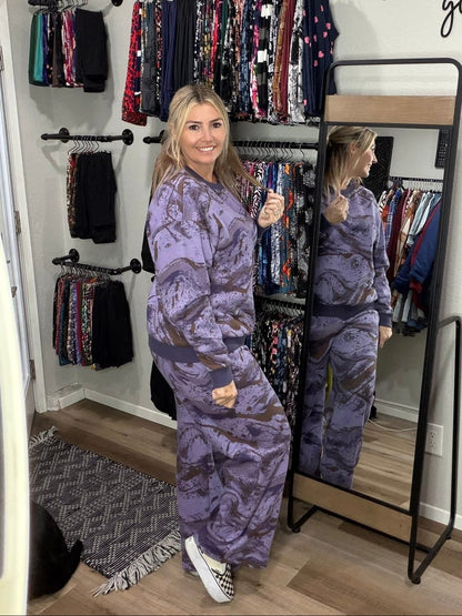 Woman wearing LuLaRoe RISE Reset  fleece crew neck pullover and RISE and Recharge sweatpants in purple marble abstract print inside boutique standing in front of a clothing rack with 'Hello Gorgeous' text.