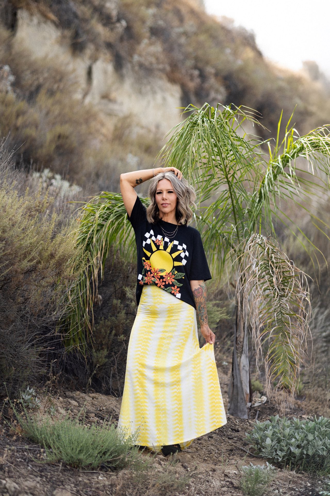 Person wearing a LuLaRoe Rivkah Top in black featuring a retro sunshine graphic with checkerboard sun rays, orange wildflowers, and vintage-inspired artwork and a yellow skirt in a natural setting. Relaxed short sleeve women's graphic top with floral and sunshine design. 