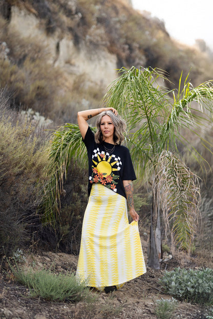 Person wearing a LuLaRoe Rivkah Top in black featuring a retro sunshine graphic with checkerboard sun rays, orange wildflowers, and vintage-inspired artwork and a yellow skirt in a natural setting. Relaxed short sleeve women's graphic top with floral and sunshine design. 