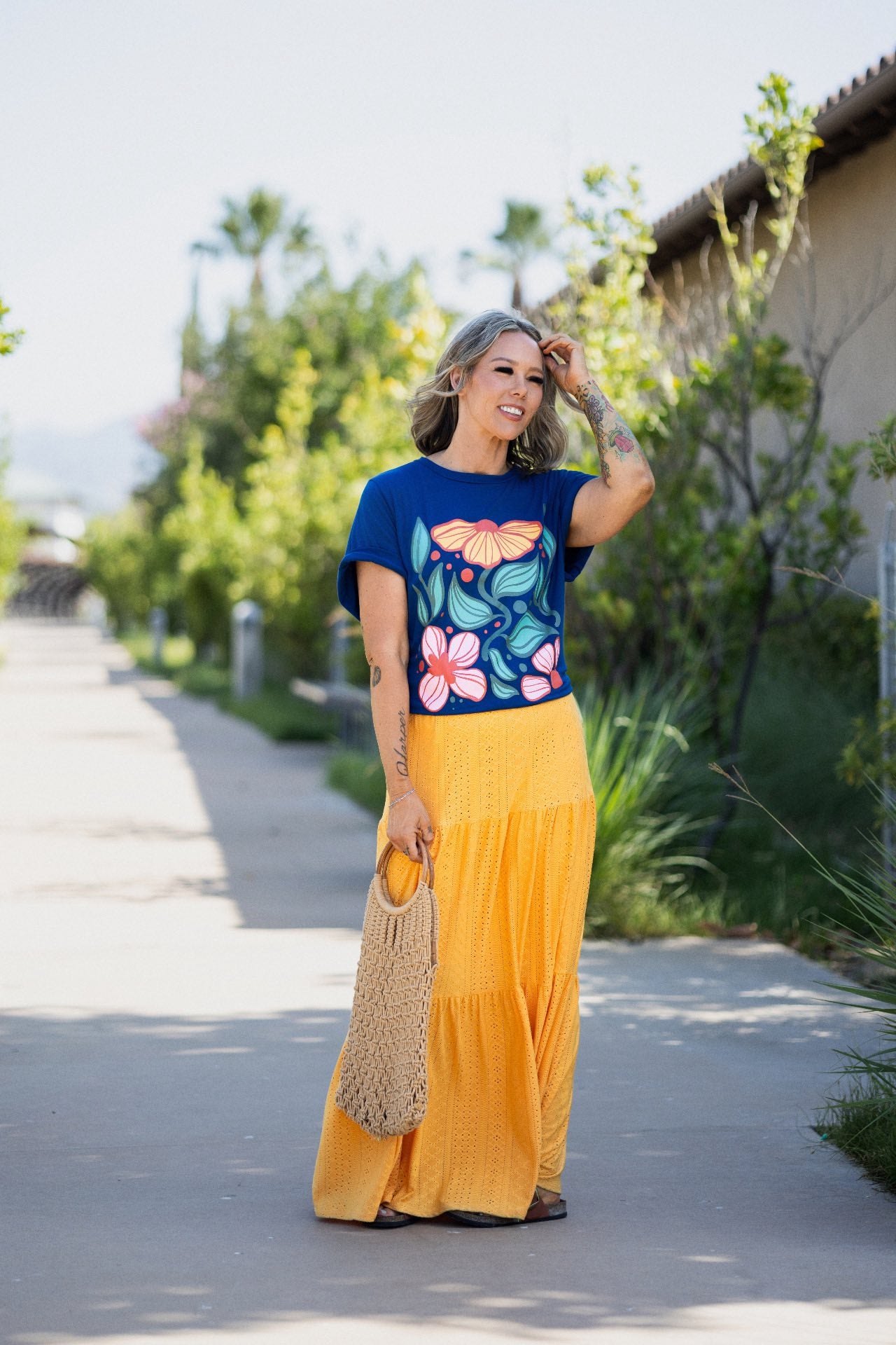 Woman wearing LuLaRoe Rivkah Top in royal blue featuring oversized retro floral artwork in pink, coral, yellow, and teal with bold botanical leaves and yellow skirt standing on a sidewalk with trees and a building in the background. Relaxed short sleeve women's graphic top with colorful floral design and artistic botanical styling.