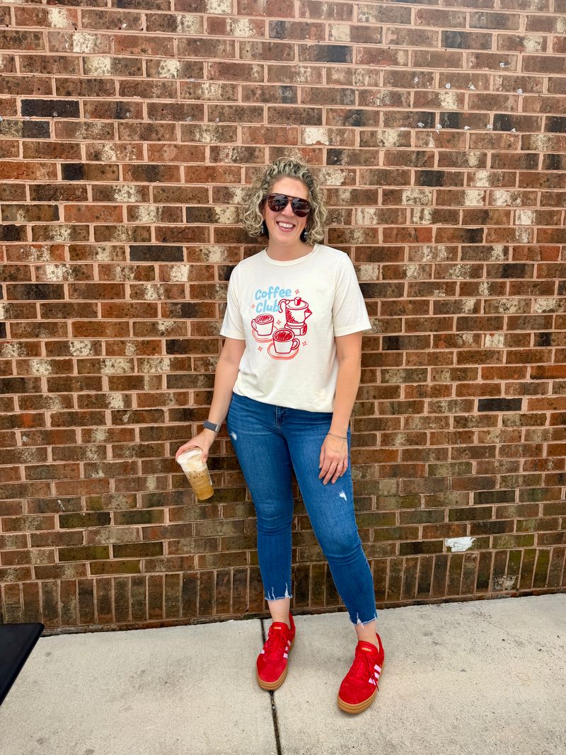 Woman wearing LuLaRoe Rivkah Top in cream featuring a Coffee Club graphic with coffee cups, moka pot, and retro typography in red and blue with  blue jeans, and red shoes, standing against a brick wall. Relaxed short sleeve women's graphic top inspired by coffee lovers, café culture, and casual everyday style