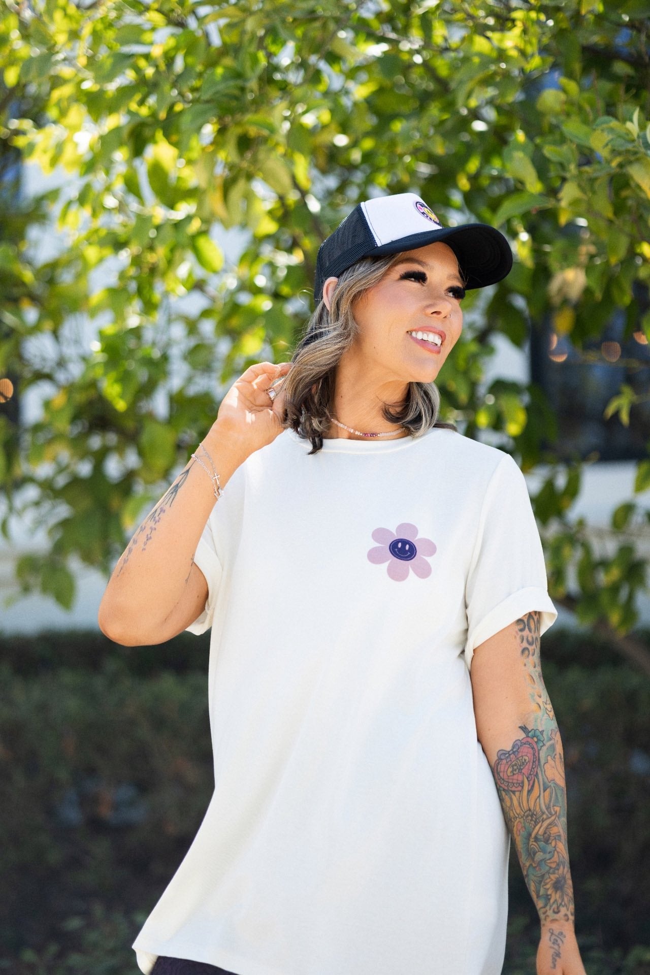 LuLaRoe Rivkah Top in white featuring a front pink smiley flower graphic and oversized back graphic reading "Have a Lovely Day" in purple and pink retro lettering with floral accents. Relaxed short sleeve women's graphic top with positive message design on model worn outdoors