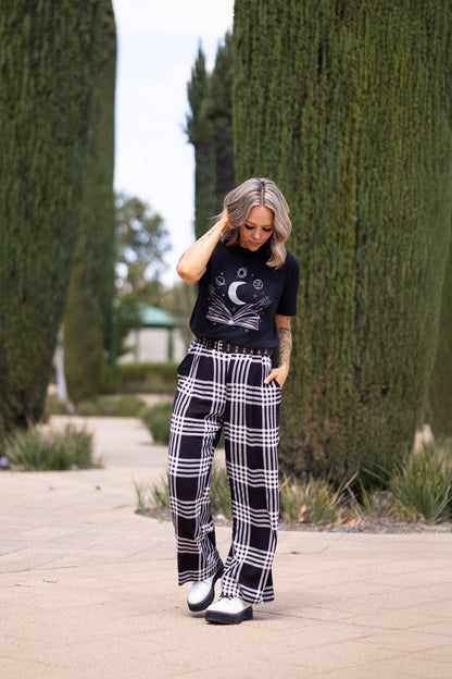 Woman wearing LuLaRoe Rivkah Top in black featuring an open book graphic with crescent moon, planets, stars, celestial symbols, and mystical artwork. Relaxed short sleeve women's graphic top with celestial reading, astrology, and book lover inspired design.and plaid pants standing on a pathway with greenery.
