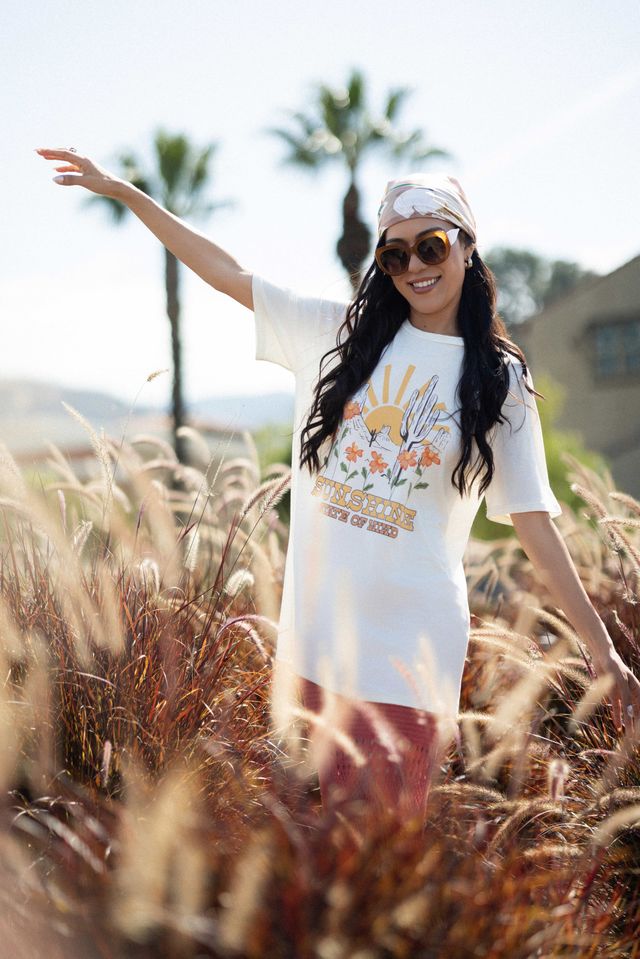 LuLaRoe Rivkah Top in white featuring a vintage-inspired sunshine graphic with wildflowers, desert landscape artwork, and retro typography. Relaxed short sleeve women's top with colorful floral details and casual everyday styling.
Woman in a white t-shirt with a graphic design, sunglasses, and cap standing in tall grass with palm trees in the background.