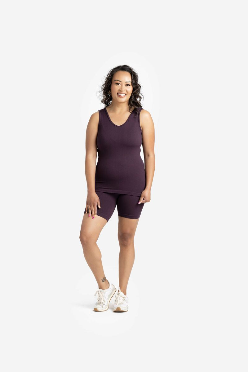 Model wearing LuLaRoe seamless biker shorts in deep plum purple with a smooth bodycon fit, high-rise waistband, and knee-length silhouette styled with a matching tank and white sneakers on a white background
