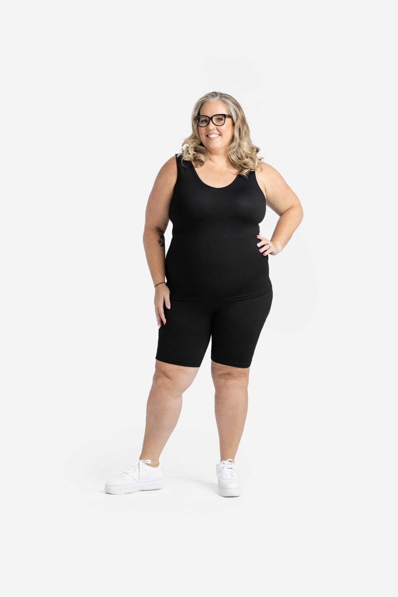 Plus-size model wearing LuLaRoe solid black seamless biker shorts with a matching black seamless tank, featuring a smooth bodycon fit, high-rise waistband, and knee-length silhouette styled with white sneakers on a neutral background
