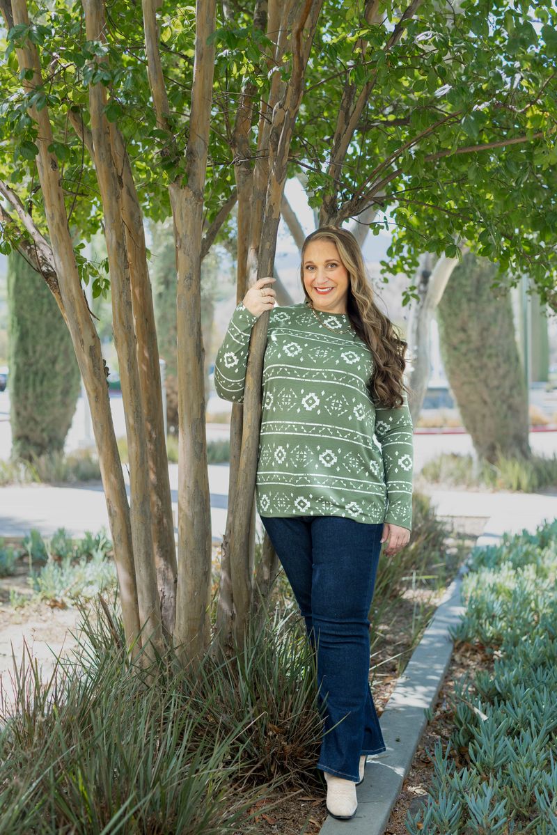 Woman wearing a green Aztec-pattern LuLaRoe Sharon Long Sleeve Top, standing outdoors near trees, styled with dark denim bootcut jeans and light boots.