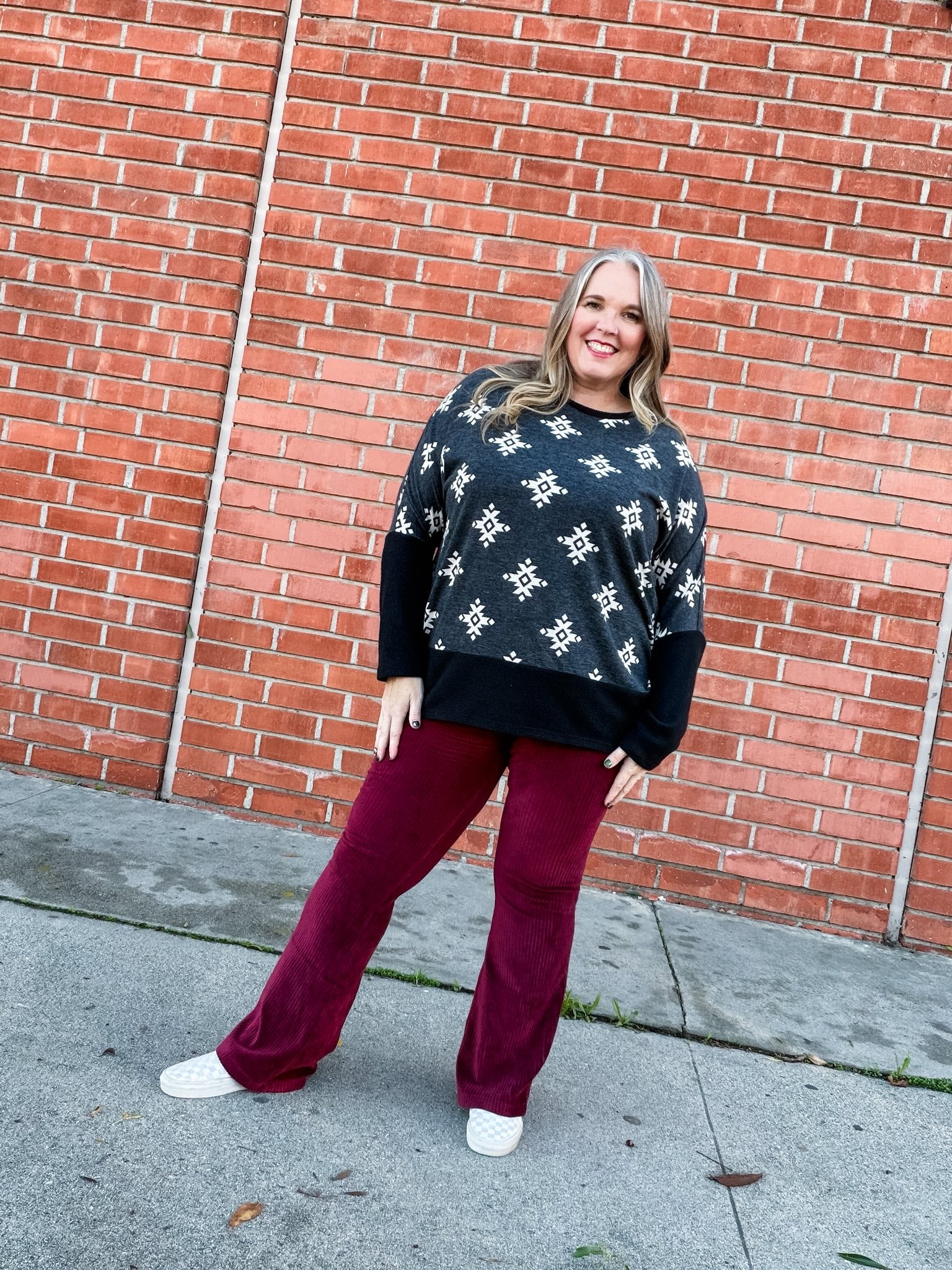 Model wearing LuLaRoe Tammy Top with black and ivory aztec print and red pants standing against a brick wall.