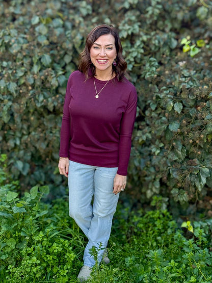 Model wearing LuLaRoe Tammy Top in deep mulberry plum solid with long sleeves standing in front of foliage