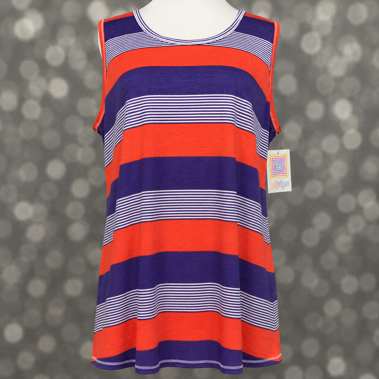 LuLaRoe tank top in Electric Orchid Stripe featuring vibrant coral and royal purple horizontal stripes accented with thin white pinstripes on a mannequin against a blurred background. Sleeveless women's tank top with a scoop neckline and relaxed fit, perfect for summer outfits, casual styling, and colorful everyday fashion. 