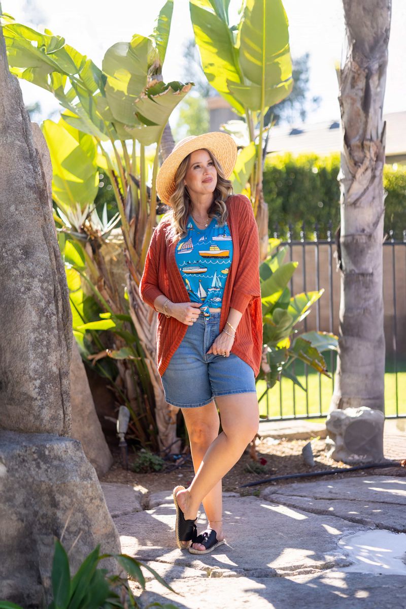 Model wearing LuLaRoe Tracy cocoon cardigan in warm rust clay orange layered over a blue graphic tee and denim shorts, featuring a relaxed draped fit and lightweight ribbed texture styled with a straw hat outdoors.