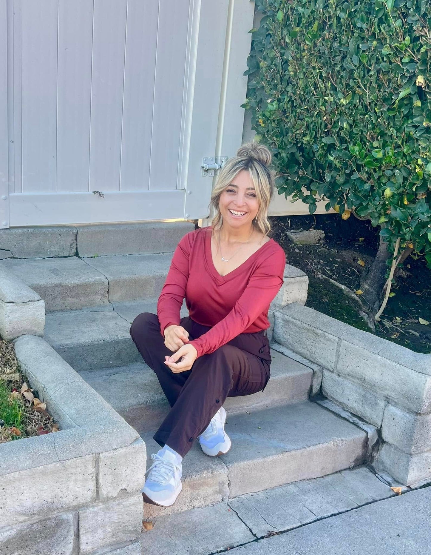 Woman wearing a LuLaRoe Vicki Top in solid brick rose with a raw-edge V-neck and long sleeves, seated outdoors on concrete steps.