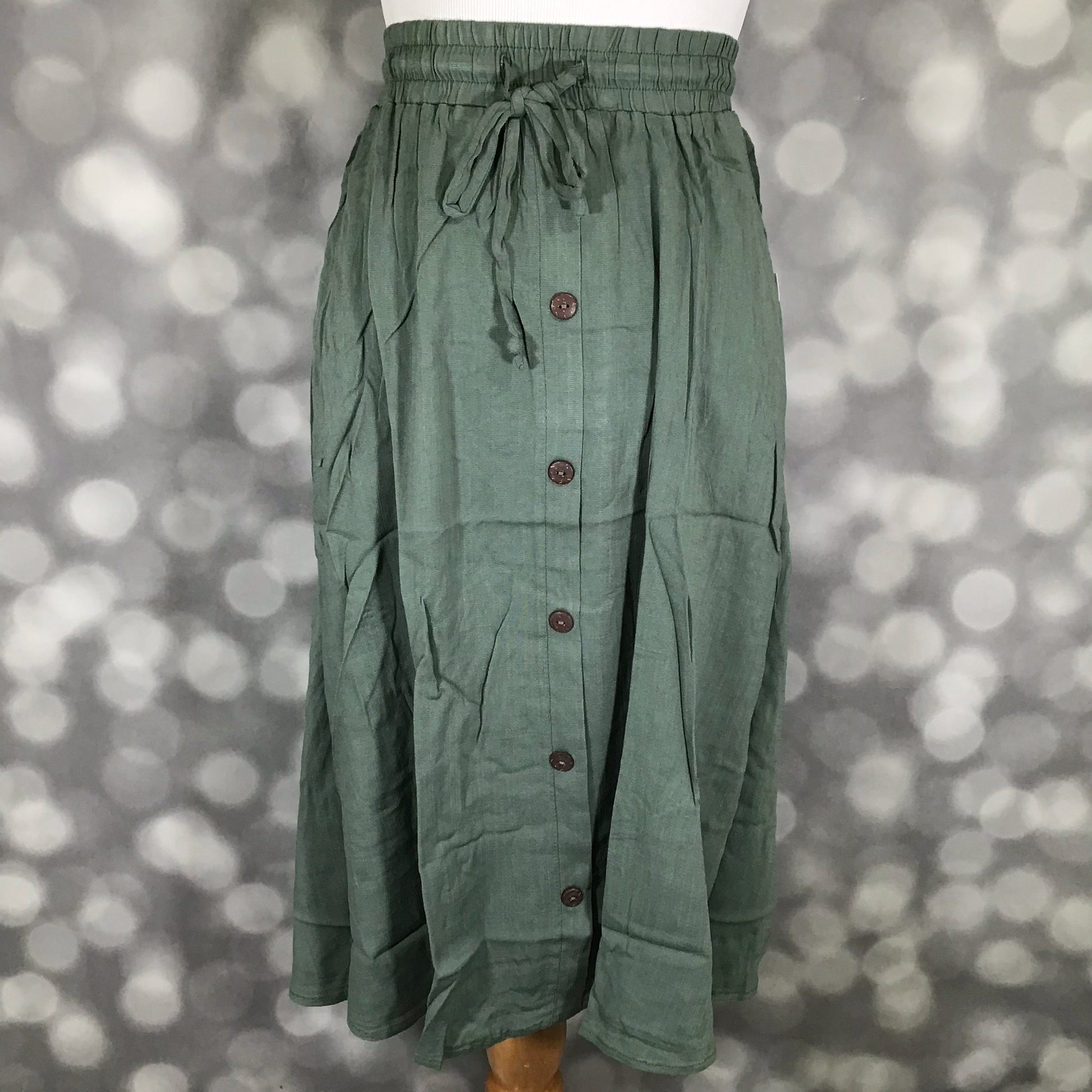 LuLaRoe Marsha Skirt Solid Sage Grove in muted green featuring a drawstring waist, button-front design, and easy everyday silhouette on a mannequin against a bokeh background