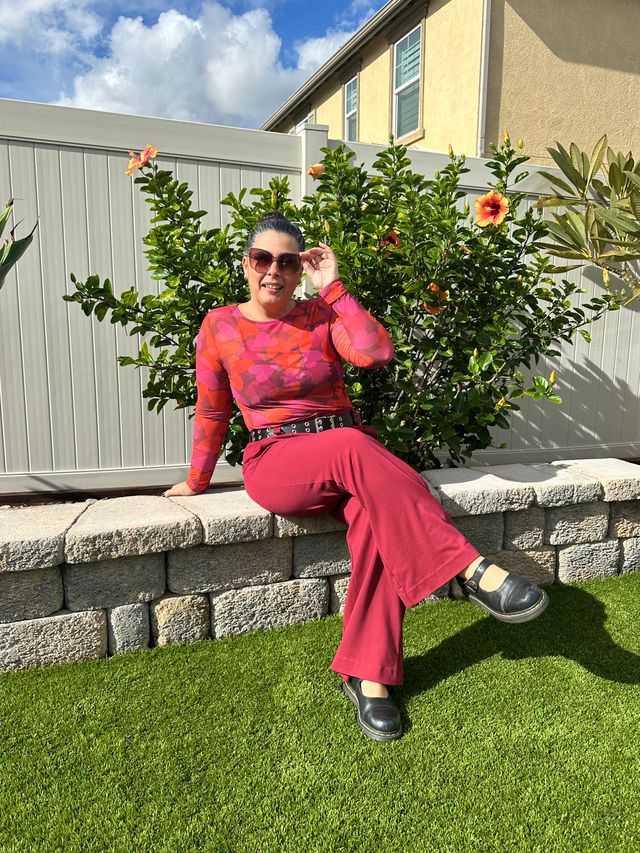 Woman wearing a LuLaRoe May long sleeve mesh top with red and pink heart pattern, fitted silhouette, lightweight semi-sheer mesh fabric styled outdoors while seated