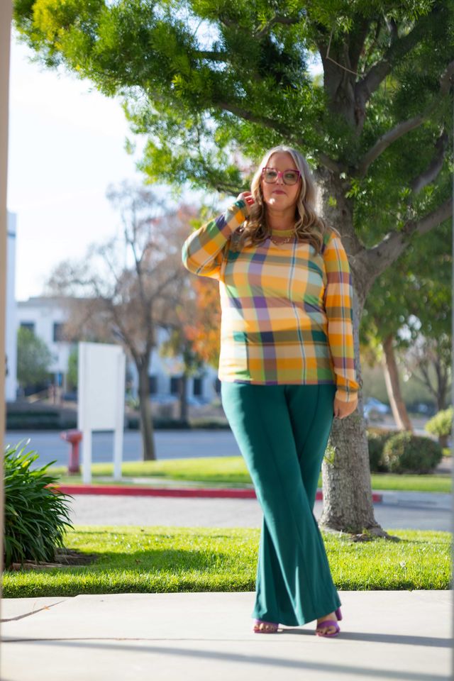 Woman wearing a LuLaRoe May long sleeve mesh top in orange and green plaid with fitted silhouette and lightweight semi-sheer mesh fabric, styled outdoors with green pants