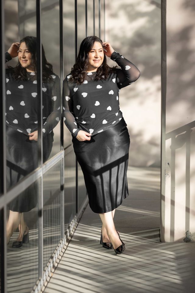 Woman wearing a LuLaRoe May long sleeve mesh top in black with scattered white heart print, fitted silhouette, semi-sheer stretch mesh fabric, styled with a black skirt