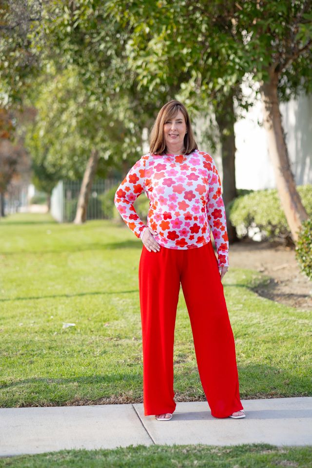 Woman wearing a LuLaRoe May long sleeve mesh top in white with red and coral floral print, fitted silhouette, lightweight semi-sheer mesh fabric styled outdoors with red pants