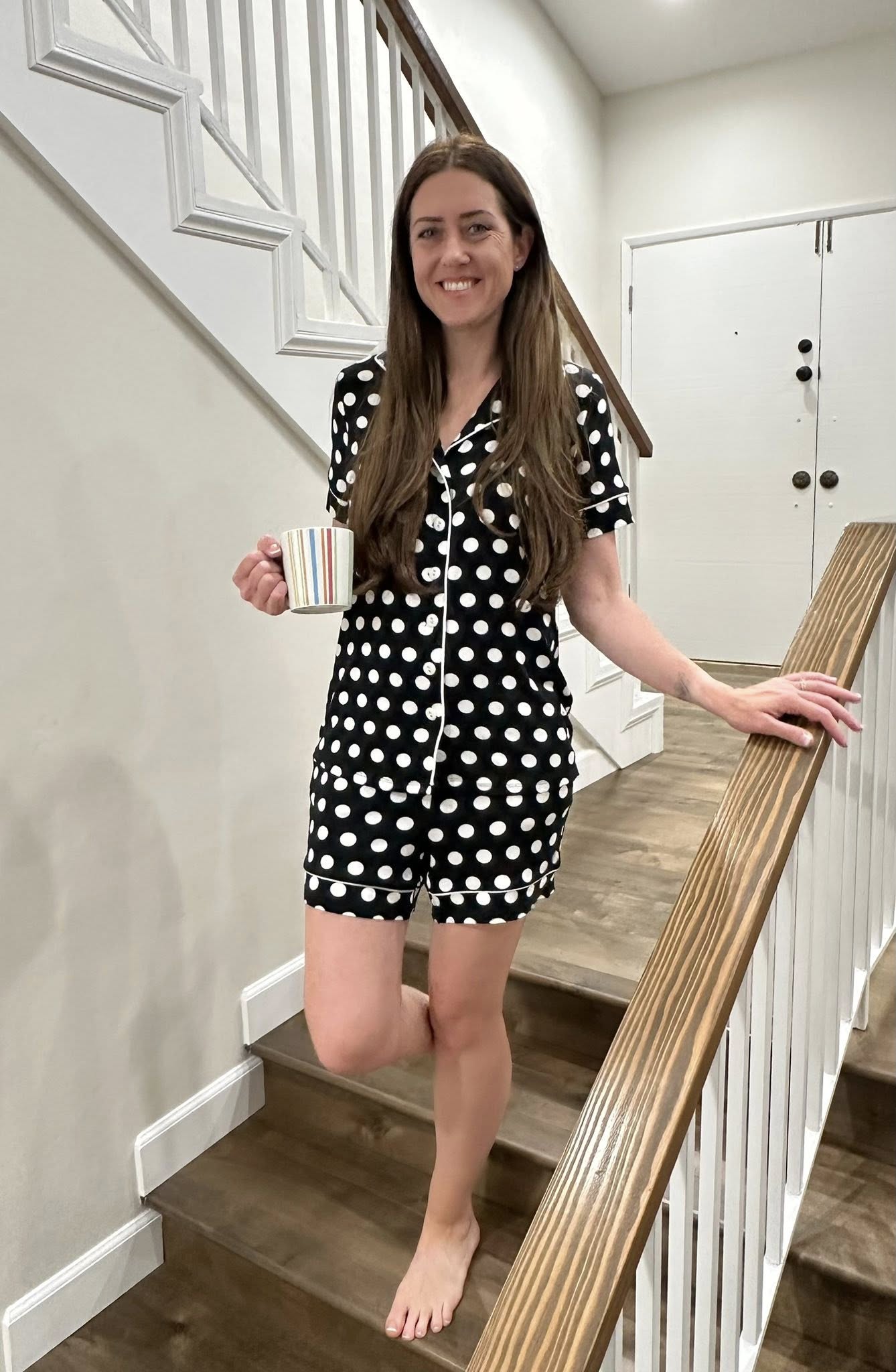 Woman wearing the LuLaRoe Candy PJ Set in a black and white polka dot print featuring a button-front short sleeve pajama top with white piping trim and matching pocket shorts in a classic polka dot sleepwear design standing on a staircase holding a mug.