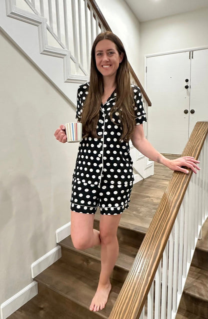 Woman wearing the LuLaRoe Candy PJ Set in a black and white polka dot print featuring a button-front short sleeve pajama top with white piping trim and matching pocket shorts in a classic polka dot sleepwear design standing on a staircase holding a mug.