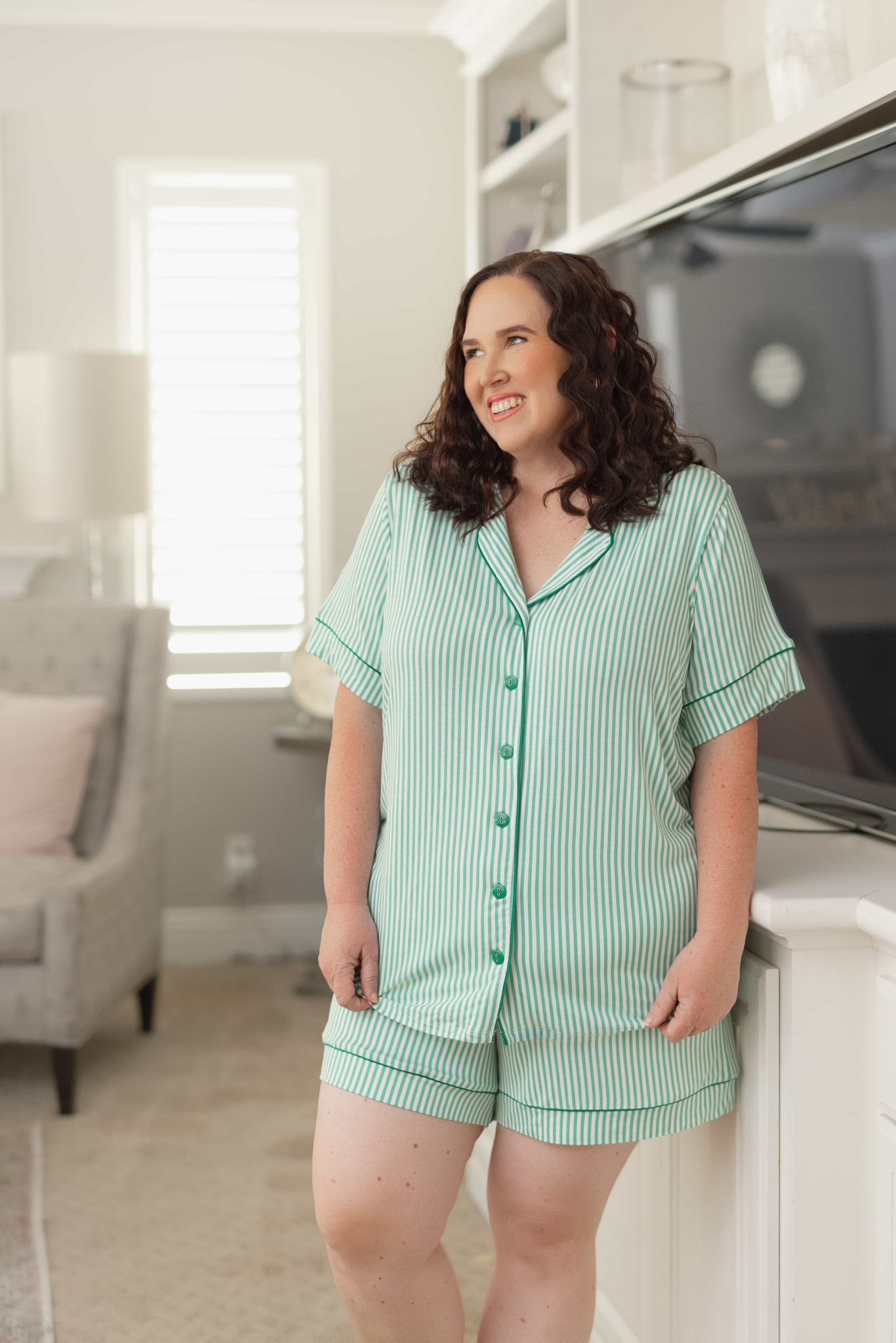 Woman wearing the LuLaRoe Candy PJ Set in a mint green and white vertical stripe print featuring a button-front short sleeve pajama top with coordinating mint piping and matching pocket shorts in a classic striped sleepwear design in a living room.