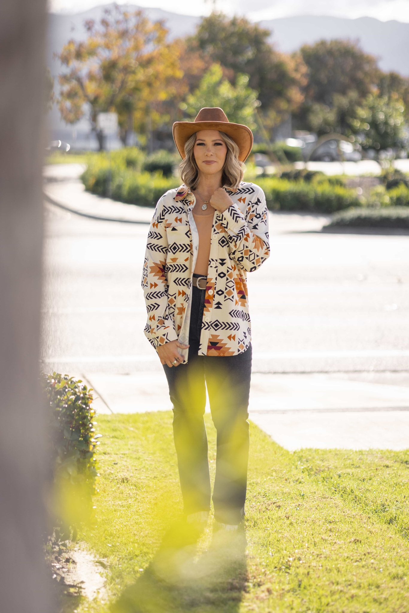 Model wearing the Myra Shacket in Canyon Weave, a cream-based southwestern geometric print shirt jacket with black and rust tones, relaxed fit, button front, and chest pockets, styled outdoors standing on grass with trees and cars in the background