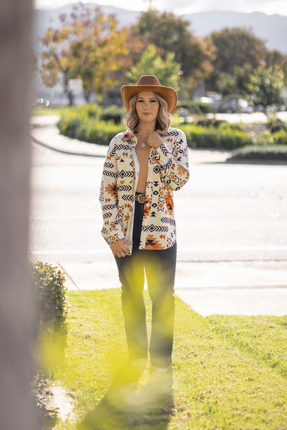 Model wearing the Myra Shacket in Canyon Weave, a cream-based southwestern geometric print shirt jacket with black and rust tones, relaxed fit, button front, and chest pockets, styled outdoors standing on grass with trees and cars in the background