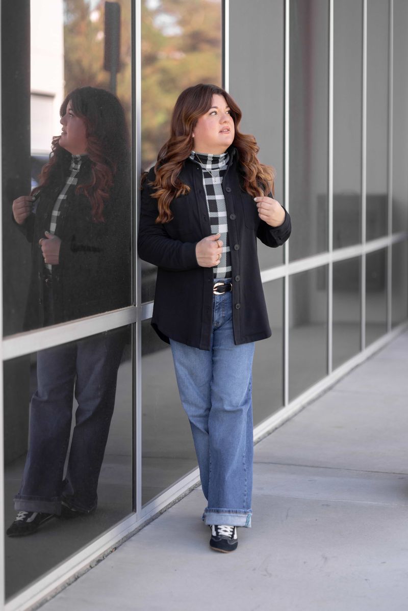 Model wearing the Myra Shacket in solid black, a relaxed-fit French terry shirt jacket with button front and chest pockets, styled with a black-and-white check top and wide-leg denim outdoors in front of a glass wall.