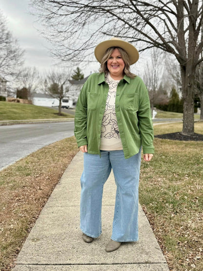 Model wearing the Myra Shacket in solid mossy olive green, a relaxed-fit French terry shirt jacket with button front and chest pockets, styled with wide-leg denim and a printed top outdoors on a sidewalk.