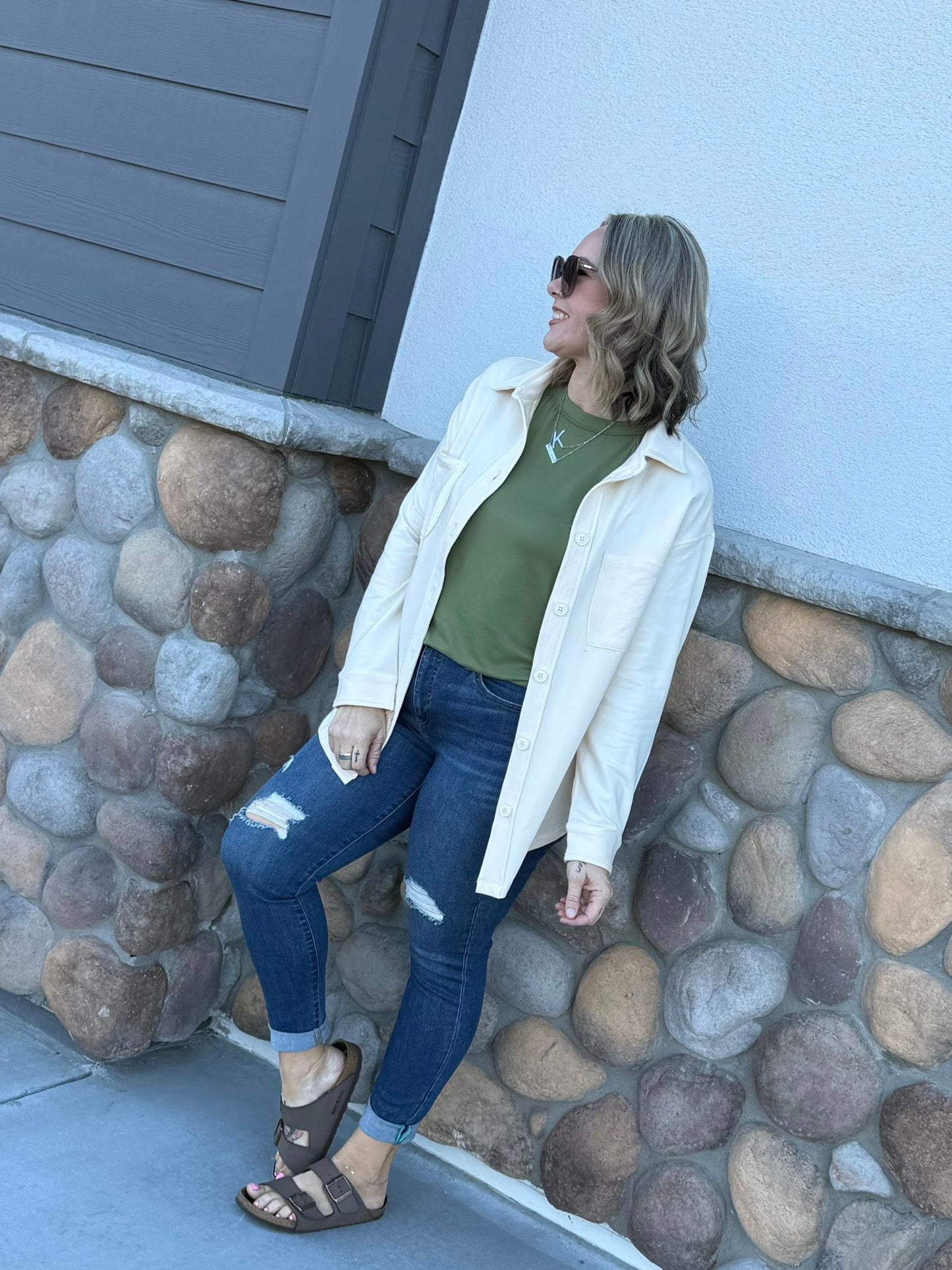 Model wearing the Myra Shacket in solid winter linen, a relaxed-fit French terry shirt jacket with button front and chest pockets, styled with a green tee and distressed denim outdoors standing against a stone wall.