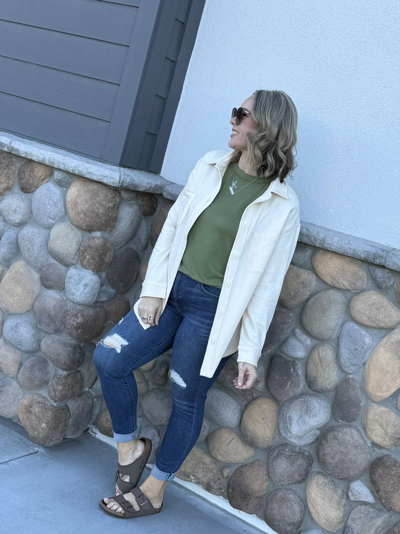 Model wearing the Myra Shacket in solid winter linen, a relaxed-fit French terry shirt jacket with button front and chest pockets, styled with a green tee and distressed denim outdoors standing against a stone wall.