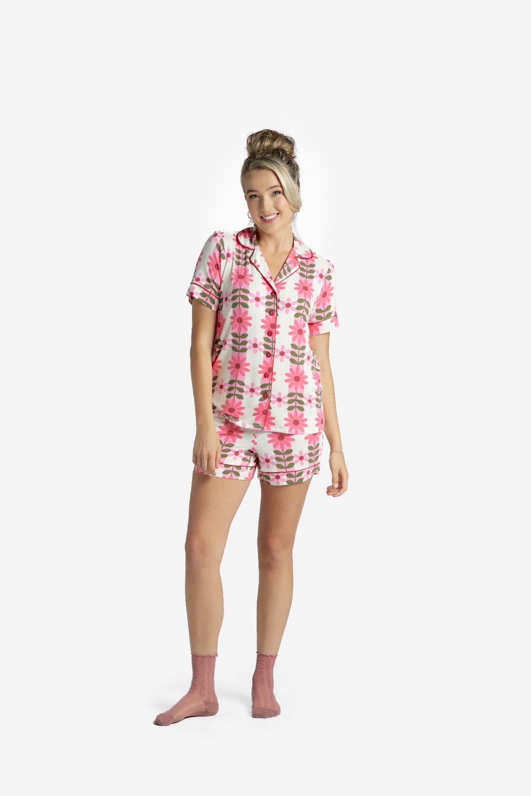 Woman wearing the LuLaRoe Candy PJ Set in a pink daisy floral print featuring bright pink flowers with olive green stems on a soft blush background, button-front pajama top, and matching pocket shorts on a white background