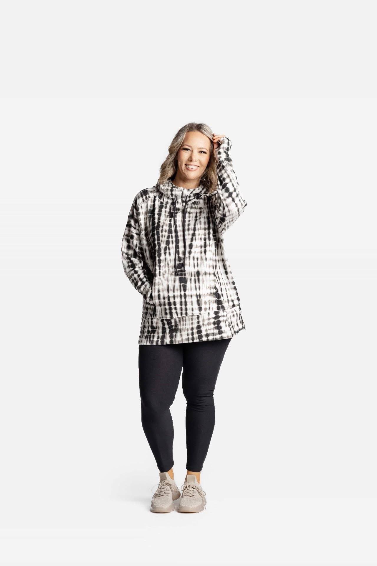 Woman wearing Regan 2.0 pullover hoodie in black and white tie-dye grid pattern with hood, drawstrings, and kangaroo pocket, styled with black leggings on a white background