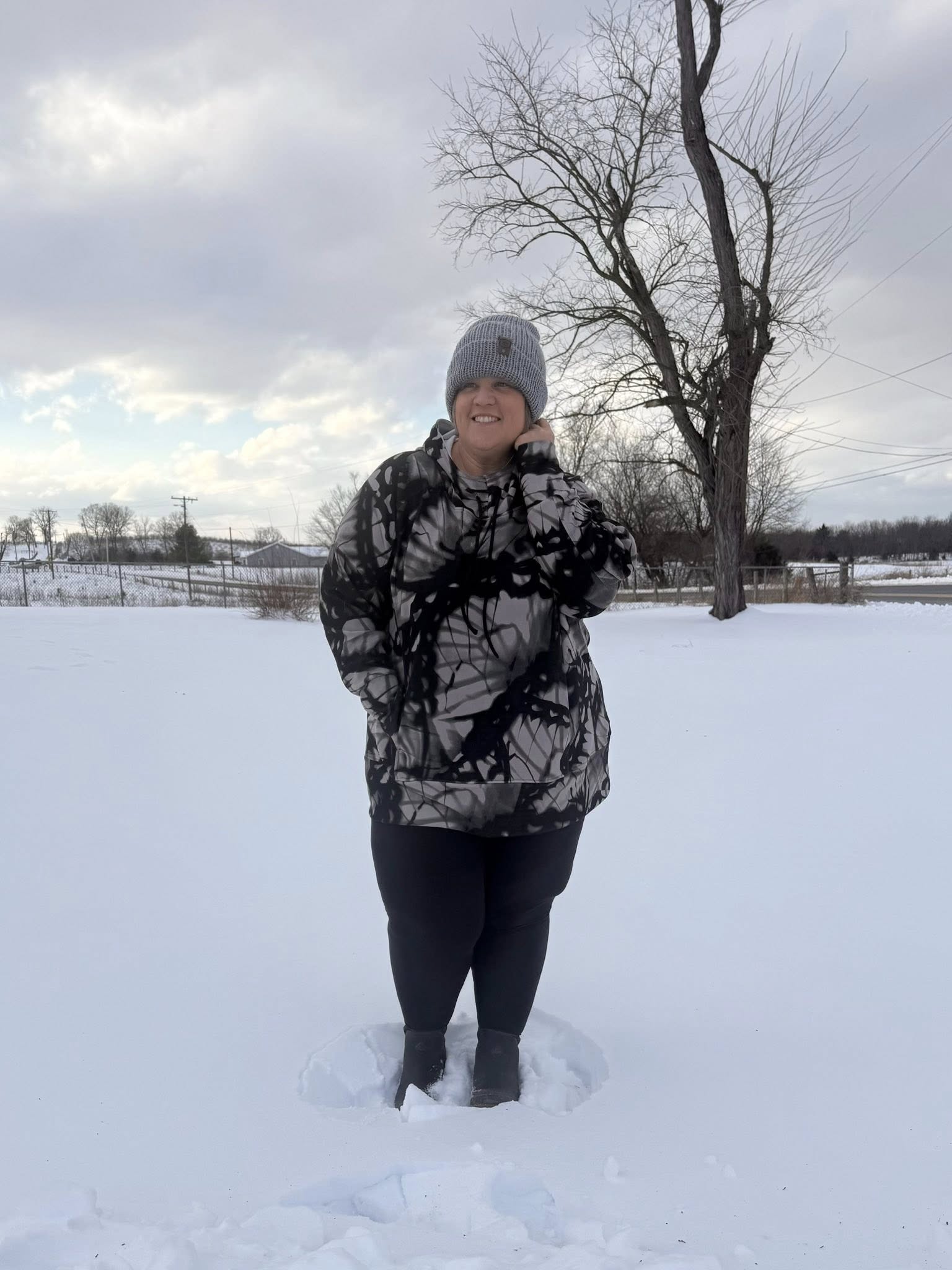 Woman wearing Regan 2.0 pullover hoodie in black and gray abstract print with hood and drawstrings, standing outdoors in snow wearing black leggings and winter boots and beanie standing in the snow.