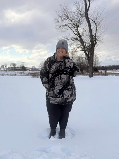 Woman wearing Regan 2.0 pullover hoodie in black and gray abstract print with hood and drawstrings, standing outdoors in snow wearing black leggings and winter boots and beanie standing in the snow.