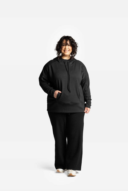 Woman modeling women’s Regan 2.0 hoodie in solid black color with relaxed fit, long sleeves, and kangaroo pocket on a white studio background