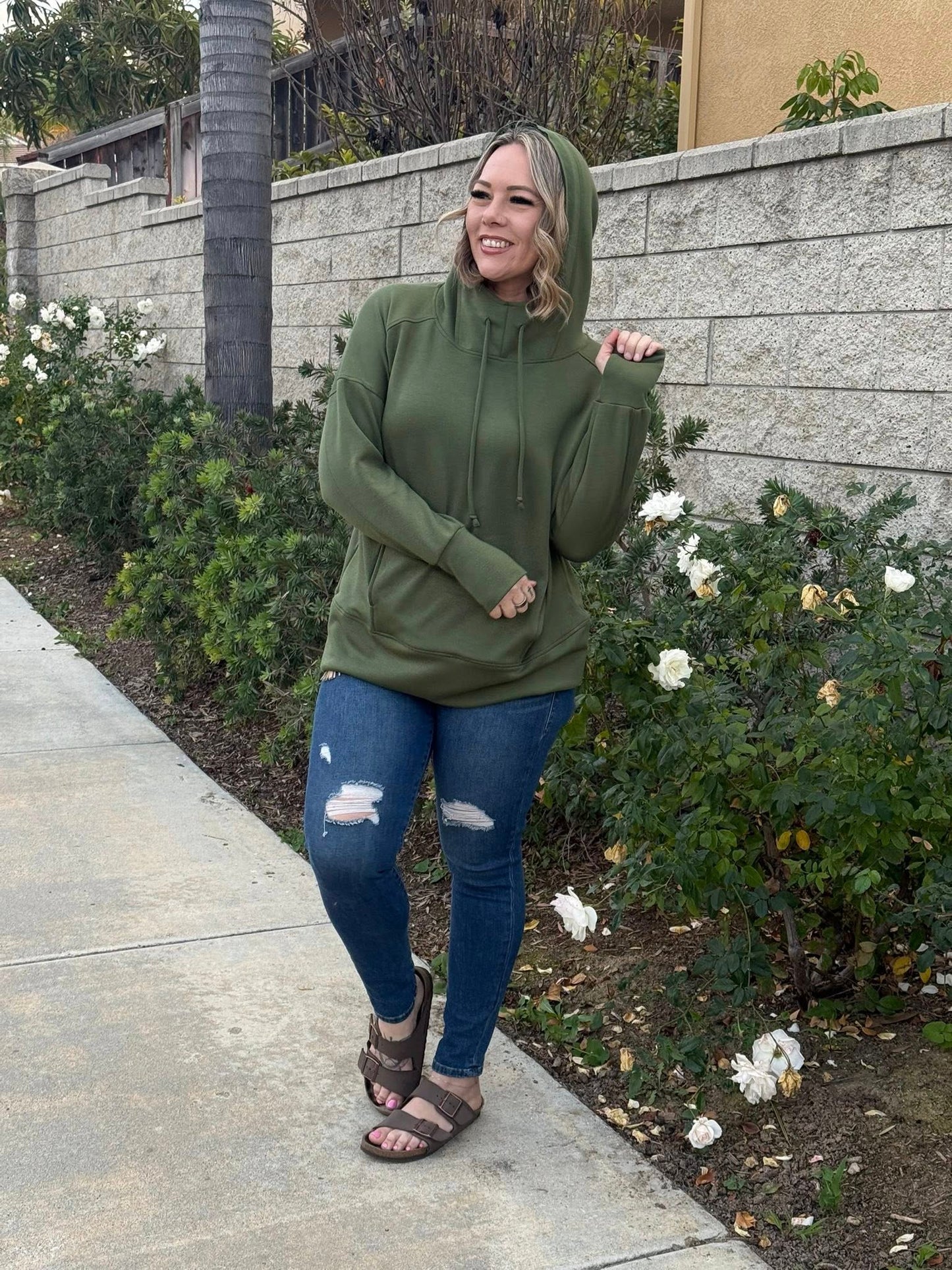 Woman wearing Regan 2.0 pullover hoodie in solid olive green with hood and drawstrings, styled with distressed blue jeans and sandals while standing on a sidewalk with a garden background
