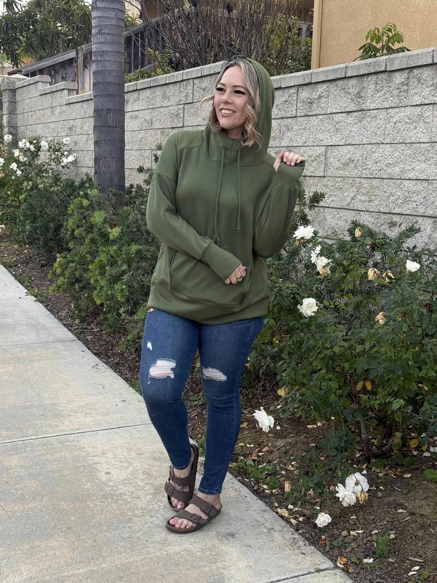 Woman wearing Regan 2.0 pullover hoodie in solid olive green with hood and drawstrings, styled with distressed blue jeans and sandals while standing on a sidewalk with a garden background