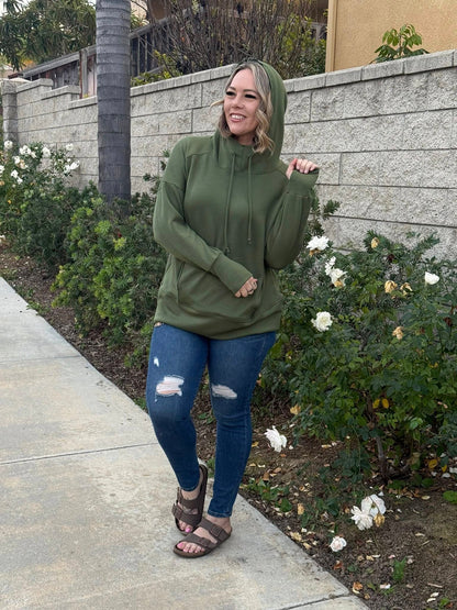 Woman wearing Regan 2.0 pullover hoodie in solid olive green with hood and drawstrings, styled with distressed blue jeans and sandals while standing on a sidewalk with a garden background