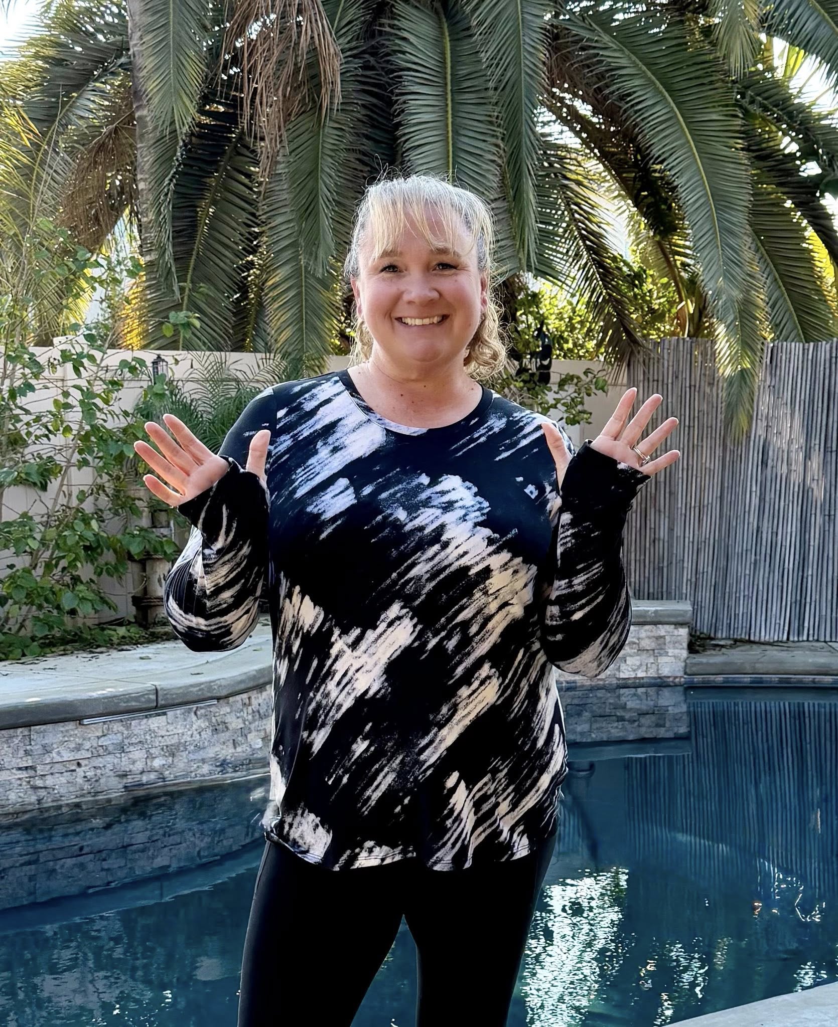 RISE Limitless Long Sleeve Top with an ivory brushstroke abstract pattern on a black background, long sleeves, and a crew neckline worn by a model outdoors by a pool with palm trees in the background