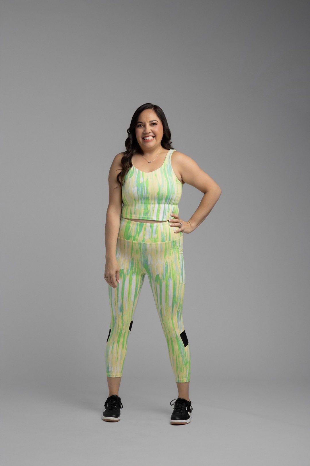 Woman modeling the LuLaRoe Rise Resilient Cami Bralette in Citrus Watercolor Drift featuring a watercolor-inspired abstract print in lime green, citrus yellow, aqua blue, and ivory with a longline athletic silhouette, paired with matching active leggings on a gray studio background.
