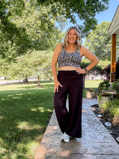 Woman modeling the LuLaRoe Rise Resilient Cami Bralette in Graphite Wildcat featuring a graphite gray and black abstract animal print longline athletic cami styled with black wide leg pants and white sneakers outdoors on a sunny walkway.