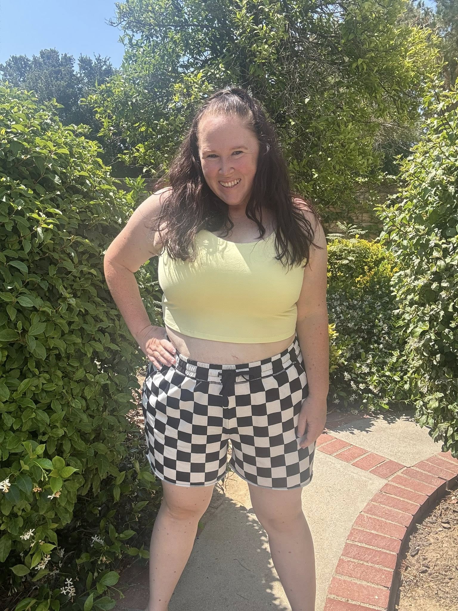 Woman modeling the LuLaRoe Rise Resilient Cami Bralette in Solid Citrus Glow, a soft pastel yellow longline athletic cami with wide straps and smooth Lotus Jersey fabric, styled outdoors with black and white checkered shorts in bright natural sunlight.