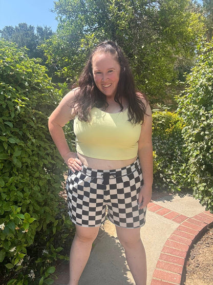 Woman modeling the LuLaRoe Rise Resilient Cami Bralette in Solid Citrus Glow, a soft pastel yellow longline athletic cami with wide straps and smooth Lotus Jersey fabric, styled outdoors with black and white checkered shorts in bright natural sunlight.