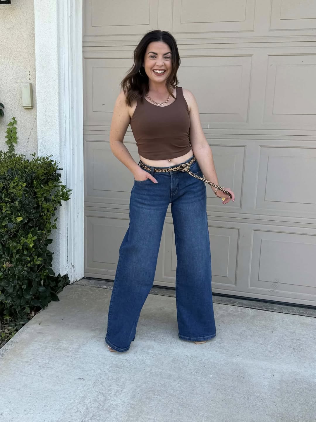 Woman modeling the LuLaRoe Rise Resilient Cami Bralette in Solid Cocoa Luxe featuring a rich brown longline athletic cami with wide straps and soft Lotus Jersey fabric, styled with wide leg denim jeans and a leopard belt outdoors in front of a garage door.
