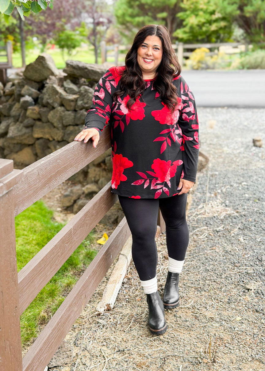 Woman wearing a LuLaRoe Sharon Long Sleeve Top in black with bold red floral print, standing outside by a wooden fence.
